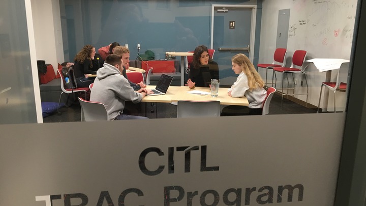 looking through a glass door with the words "CITL TRAC Program" etched on it. two groups of student sit at 2 tables, one in the foreground and one in the background. each group appears to be working collaboratively on a project. 
